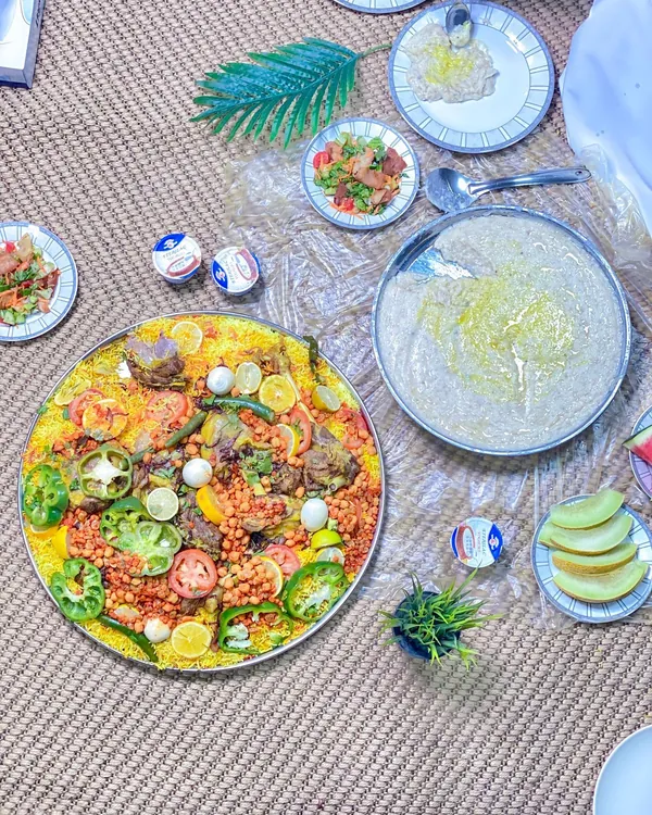 Overhead view of a large rice platter with meat, tomatoes, peppers and chickpeas, beside a platter of creamy porridge, salads and melon